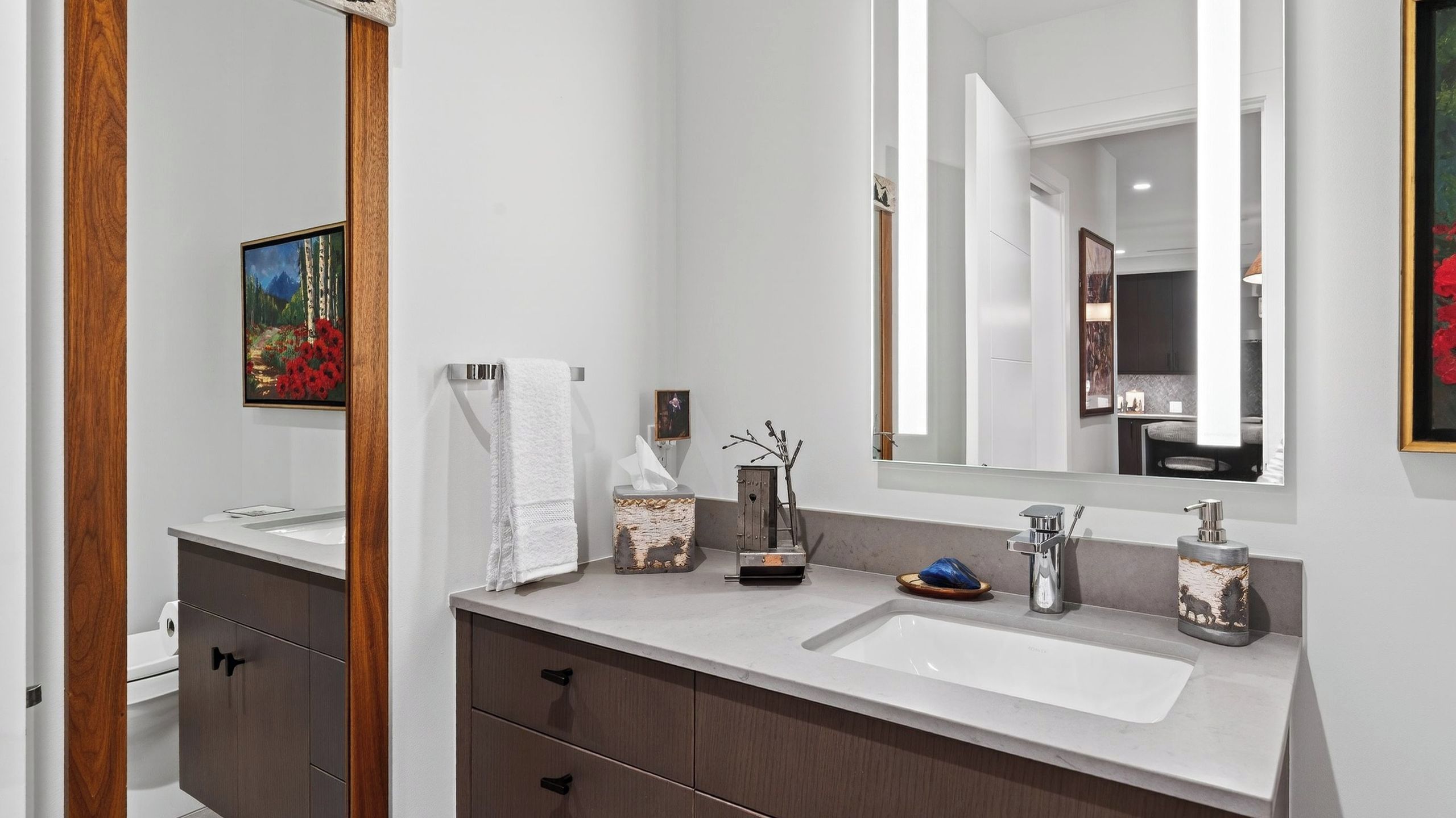 A modern bathroom with a large mirror, a white sink, and a wooden vanity.
