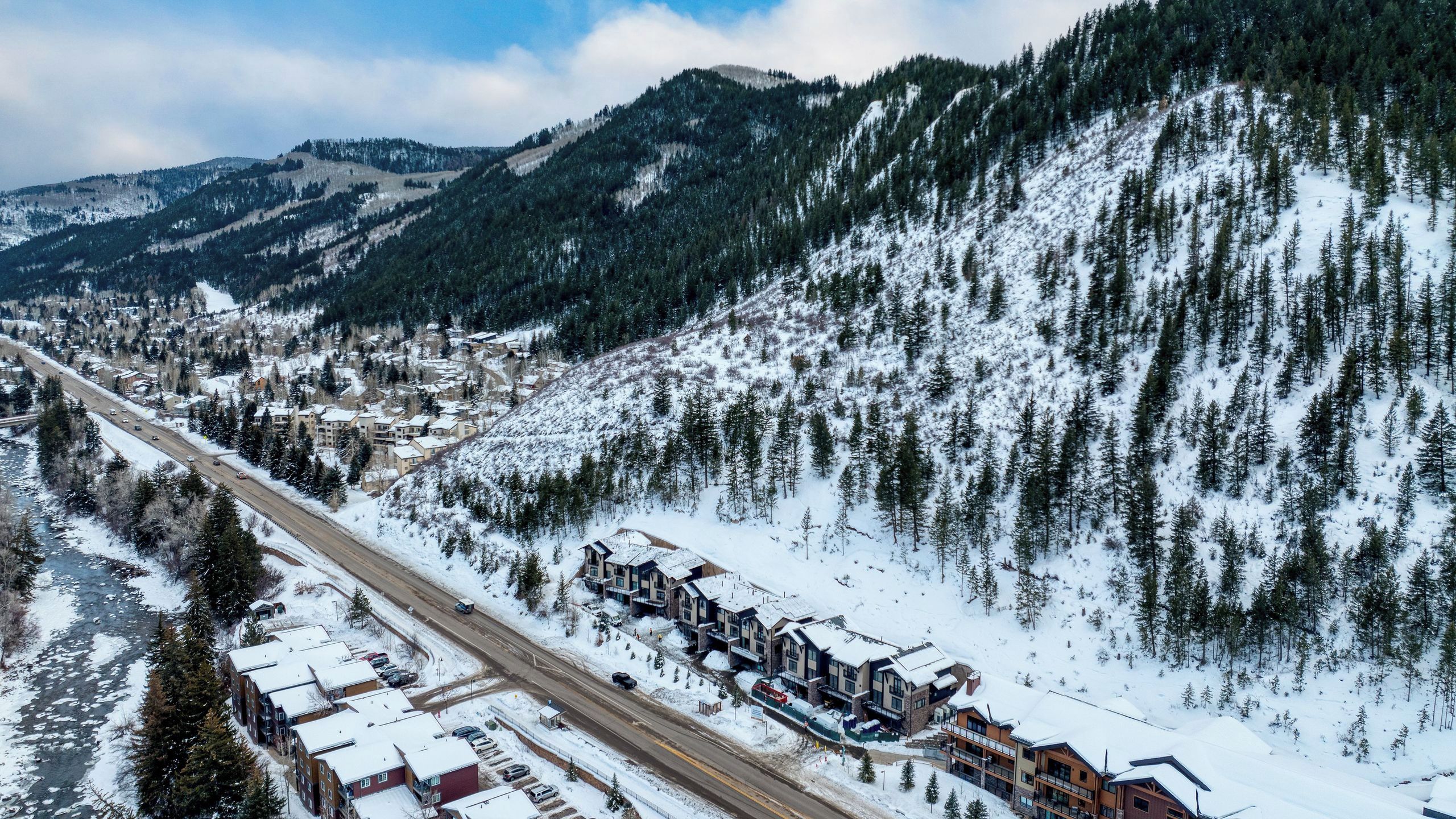 A snow-covered mountain town with a river, a highway, and a row of multi-story buildings with balconies.