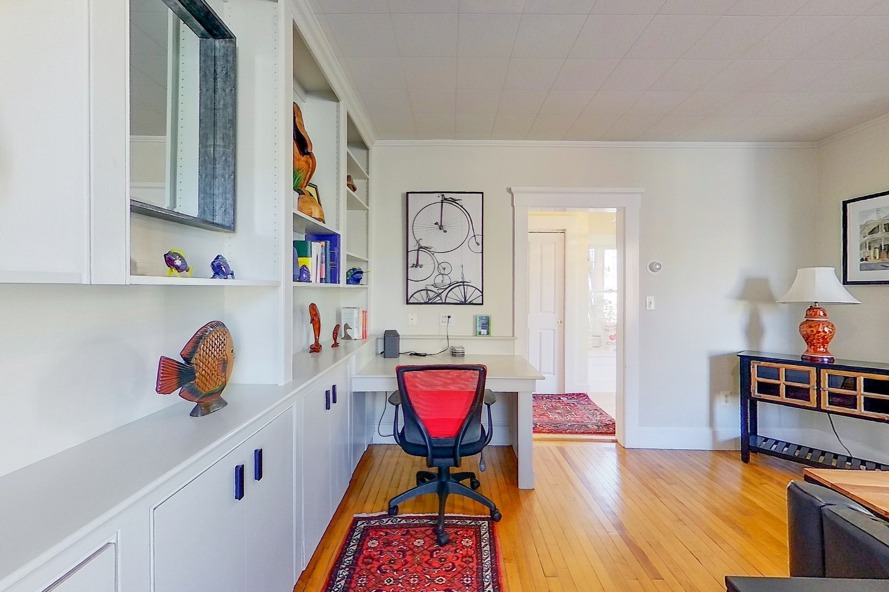 A modern living room with a white desk, a red office chair, a patterned rug, and a bookshelf with decorative items.