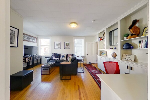 A spacious living room with a black sofa, a wooden coffee table, and a red chair. There is a television on the left and a bookshelf with various items on the right.