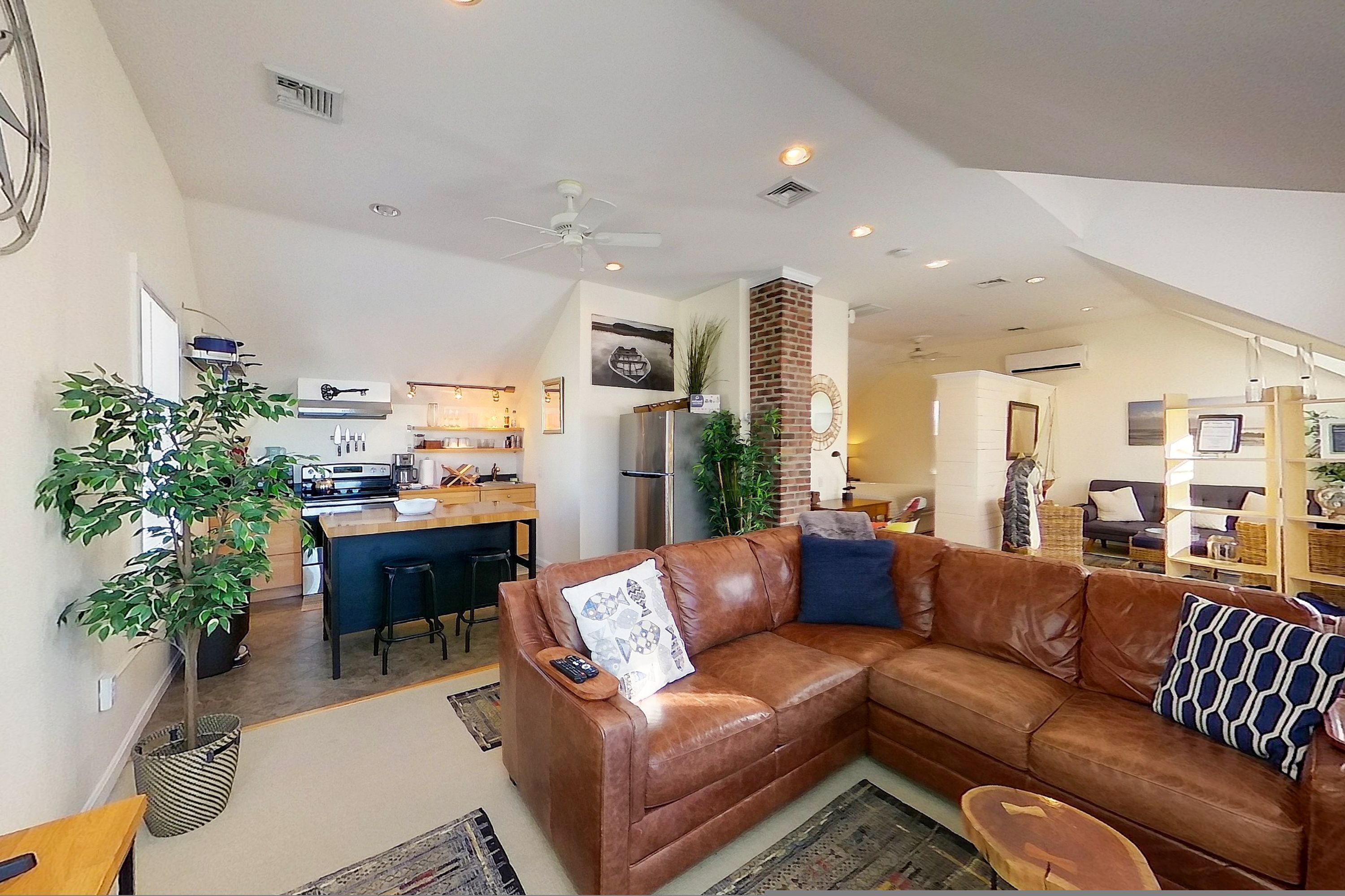 A modern living room with a brown leather sofa, a dining area with a kitchen in the background, and a staircase leading to another level.