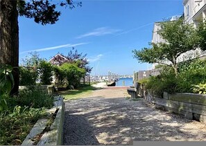 Un chemin de gravier mène à une marina où sont amarrés des bateaux, le tout étant entouré de verdure et de bâtiments.