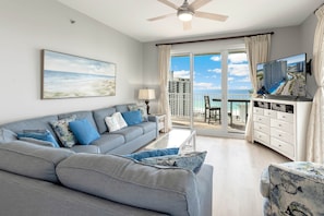 A living room with a grey sofa, a television, and a balcony with a view of the ocean and buildings.
