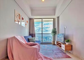 A modern living room with a large window, a sofa, a TV, and a plant. - RIA By SanLiving - 3BR - Hublife Mall (Jakarta)