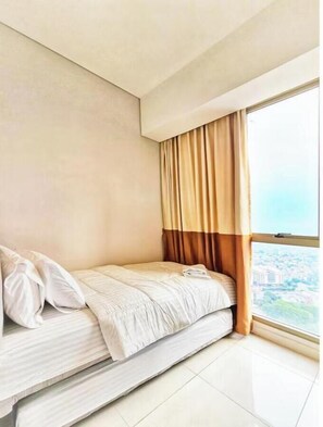 A small, single-bed room with a city view through a large window, a curtain, and a plain wall. - RIA By SanLiving - 3BR - Hublife Mall (Jakarta)