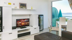 Ett modernt vardagsrum med en platt-TV, vita skåp och en glasdörr som leder ut till en balkong med utsikt över havet.