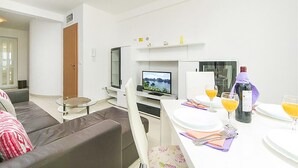 Ett modernt vardagsrum med en platt-TV, ett matbord dukat med tallrikar, glas och en flaska, och en soffa med en kudde.
