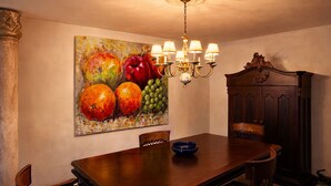 Een eetkamer met een houten tafel, stoelen, een kroonluchter en een schilderij van fruit aan de muur.