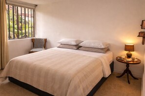 Ein ordentlich bezogenes Bett mit einer beigefarbenen Tagesdecke, ein Nachttisch mit einer Lampe und ein Sessel.