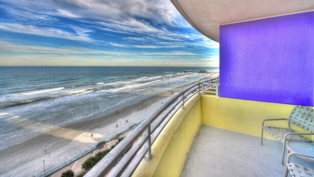 Ein Balkon mit Blick auf Strand und Meer, eine violette Wand und eine Sitzgelegenheit mit Metallstühlen.