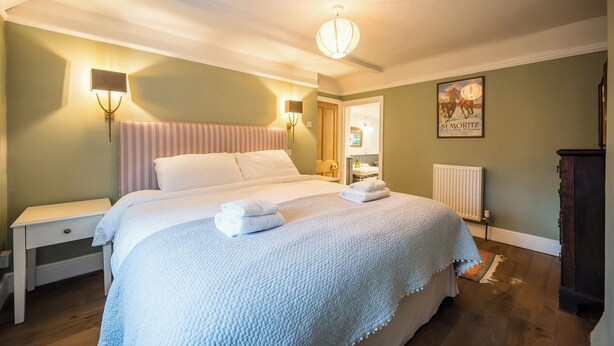 Un lit bien fait, avec du linge de lit blanc et deux serviettes pliées, une table de chevet, une lampe fixée au mur et un radiateur.