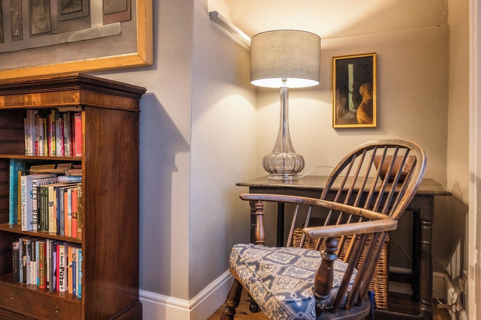 Un coin d’une pièce avec une bibliothèque en bois remplie de livres, un fauteuil à motifs, un grand lampadaire et un tableau encadré accroché au mur.