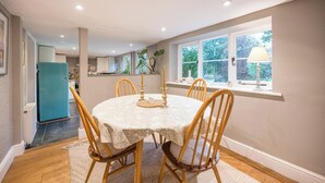 Un coin repas avec une table ronde, quatre chaises en bois, une nappe blanche et une fenêtre donnant sur un paysage verdoyant.