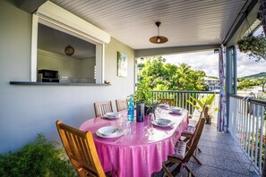 Un espacio exterior cubierto para comer con una mesa dispuesta para cuatro personas, sillas alrededor y un microondas visible a través de una ventana abierta.