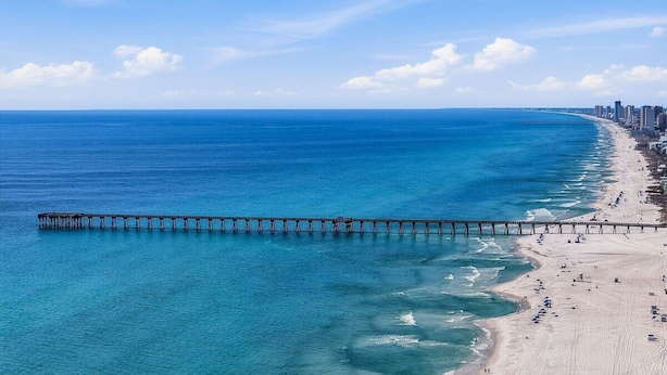 Una playa costera con un largo muelle que se adentra en el océano, una orilla arenosa y un horizonte urbano distante.