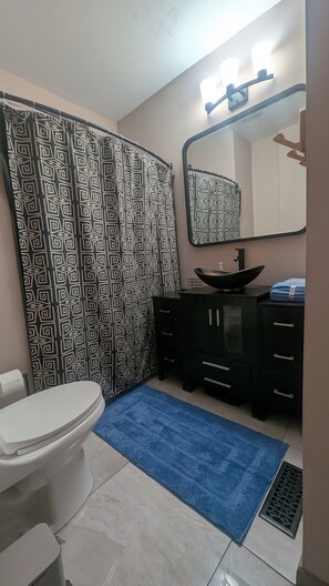 A bathroom with a toilet, a sink with a black basin, a mirror above the sink, and a shower curtain with a geometric pattern. - Craftsman house near downtown KC and MCI airport (Kansas City)
