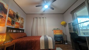 A bedroom with a bed, a desk, a chair, a ceiling fan, and a window with a view.