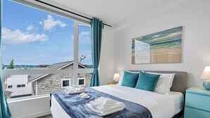 A hotel room with a large window, a bed with blue bedding, a painting of a beach, and a view of buildings outside.