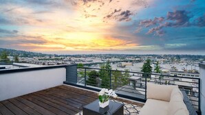 A rooftop terrace with a wooden deck, a seating area with a sofa and a table, and a cityscape view during sunset.