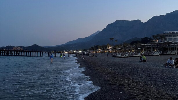 A beach with a pier extending into the water, people walking along the shore, and mountains in the background. - ESG • 5-Min Walk to Göynük Beach • Mountain View Terrace Studio (Goynuk)