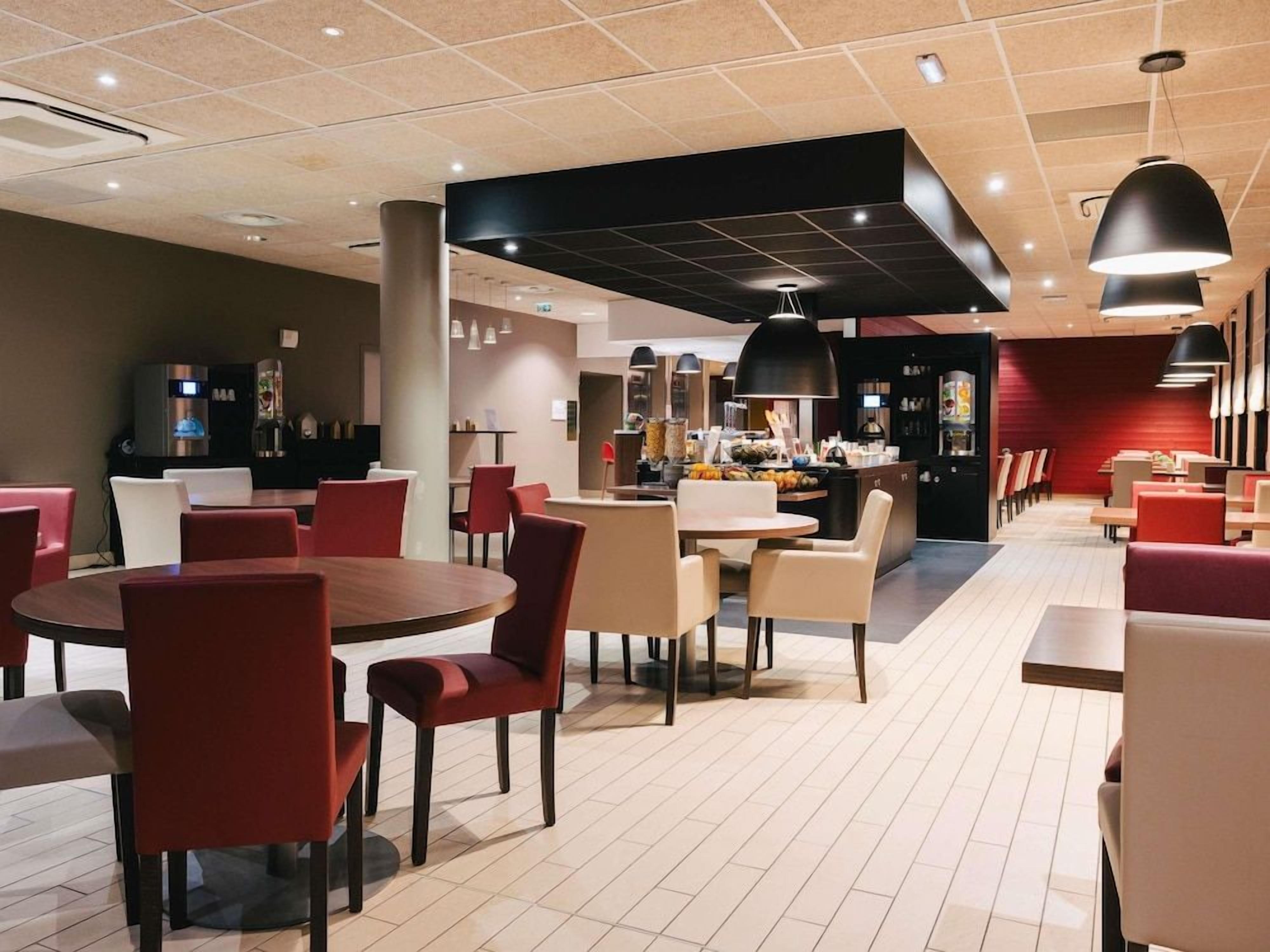 A modern restaurant interior with round tables and chairs, a bar area, and a kitchen visible in the background.