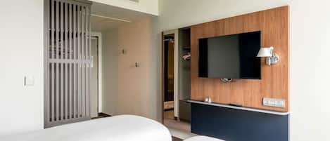 A hotel room with a bed, a TV mounted on a wooden panel, and a door with a vertical metal grille.
