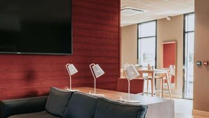 A modern interior with a dark grey sofa, a flat-screen TV mounted on a red wall, and a wooden desk with two white lamps. - Campanile Rouen Mermoz (Rouen)