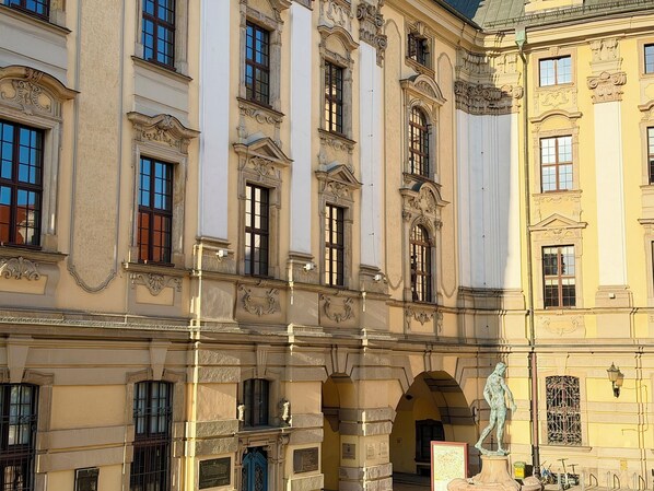 Un edificio histórico con arquitectura clásica, que presenta columnas ornamentadas, ventanas amplias y una estatua en el patio.