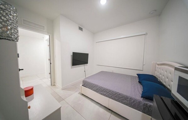Ein Schlafzimmer mit einem Bett, einem an der Wand montierten Fernseher, einer Tür und einem kleinen Tisch mit einem roten Gegenstand darauf.
