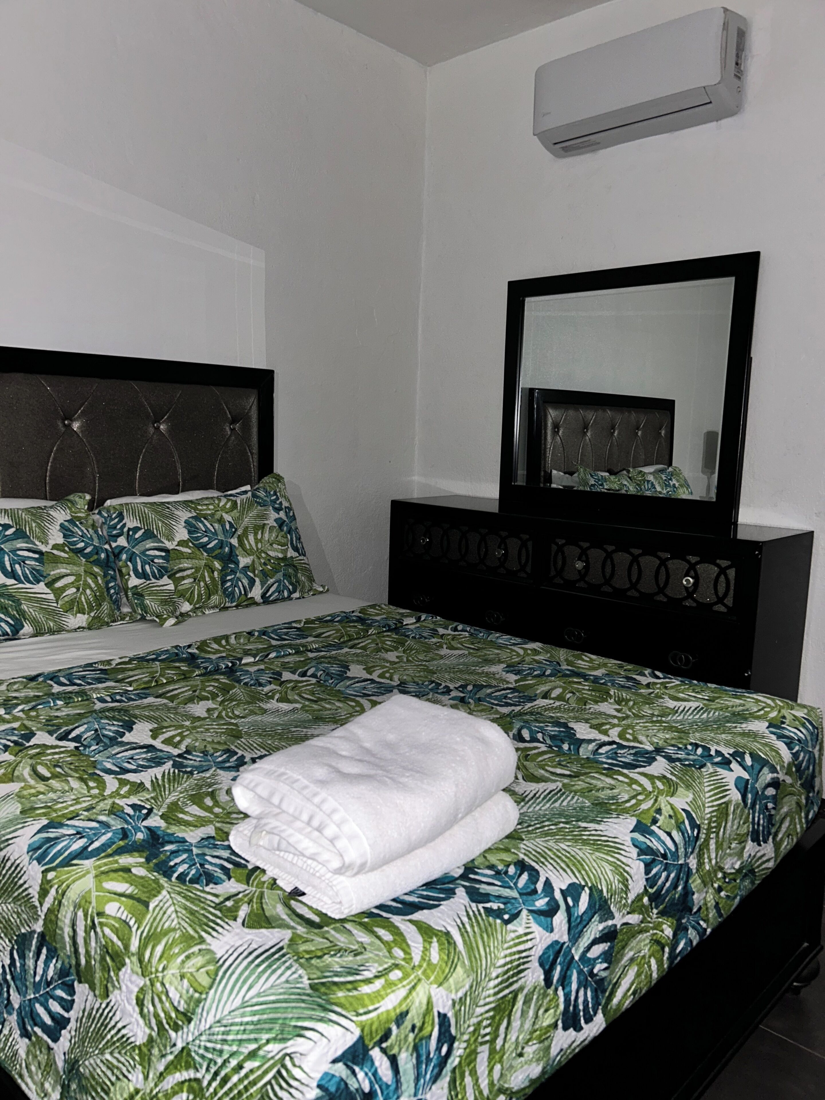 A neatly made bed with a tropical leaf pattern bedspread, a black headboard, and a large mirror above it.