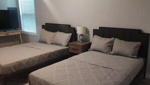 Ein Zimmer mit zwei Betten, jeweils mit Kopfstütze, einem Nachttisch und einer Lampe sowie einem Fernseher auf einem Standfuß.
