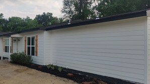 Ein eingeschossiges Haus mit Veranda, Garage und einem kleinen Vorgarten mit Buschwerk und Pflanzen.