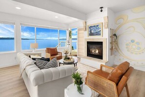 A spacious living room with a fireplace, large windows overlooking a body of water, and a view of the sky.
