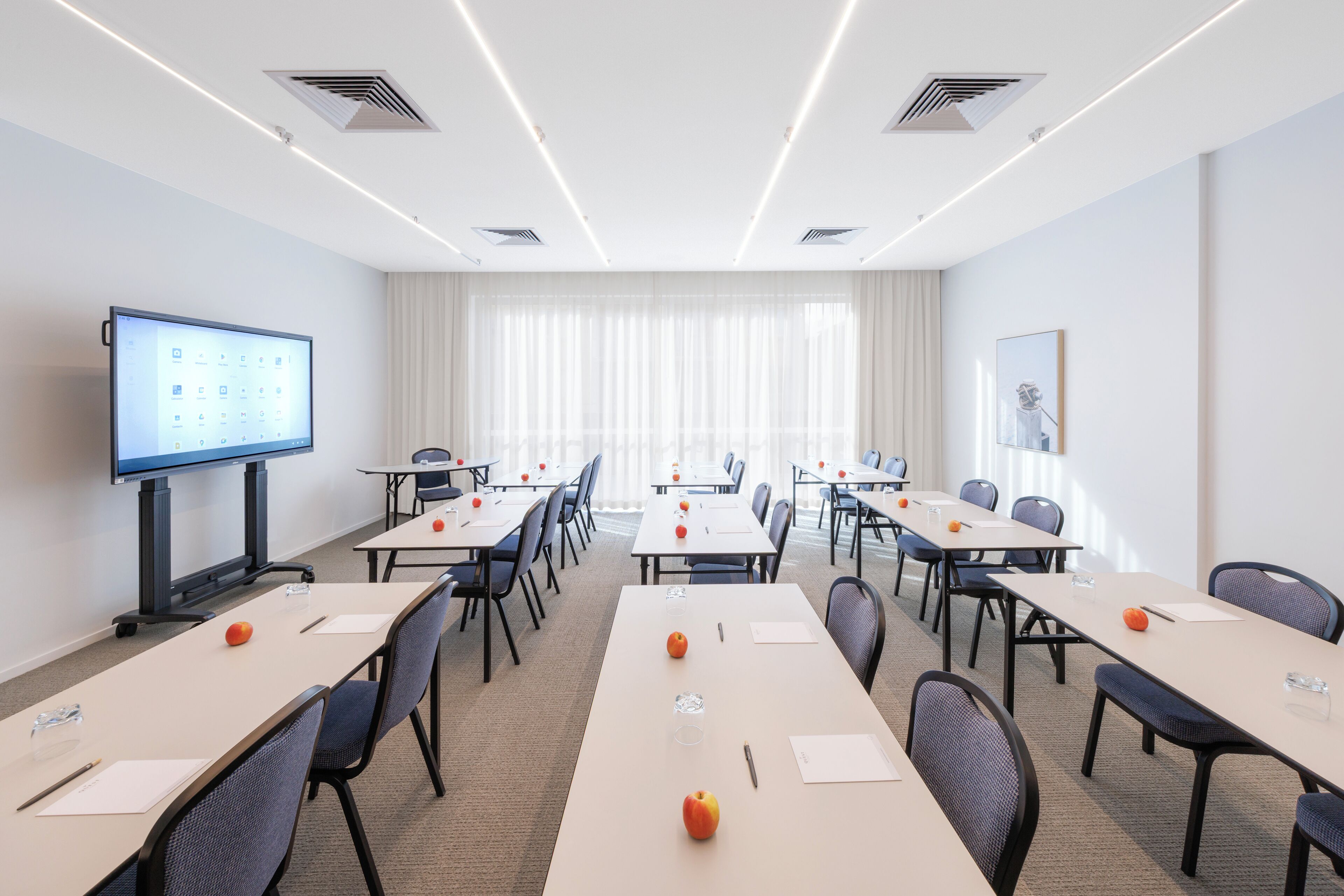 Un salón de conferencias con una pantalla grande, varias mesas, sillas y un ventanal con cortinas.