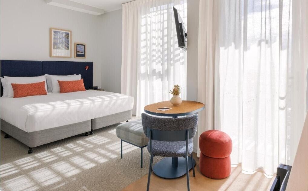 A hotel room with a bed, a round table, a chair, and a red ottoman.