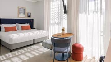 A hotel room with a bed, a round table, a chair, and a red ottoman.