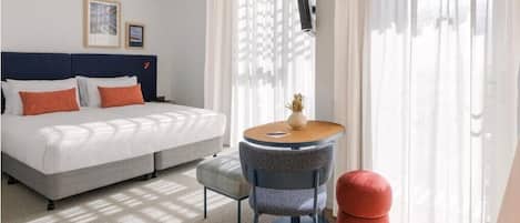 A hotel room with a bed, a round table, a chair, and a red ottoman.