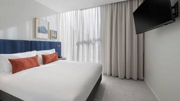 A hotel room with a large bed, a flat-screen TV mounted on the wall, and a window with sheer curtains.