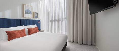 Habitación de hotel con una cama grande, un televisor de pantalla plana en la pared y una ventana con cortinas translúcidas.