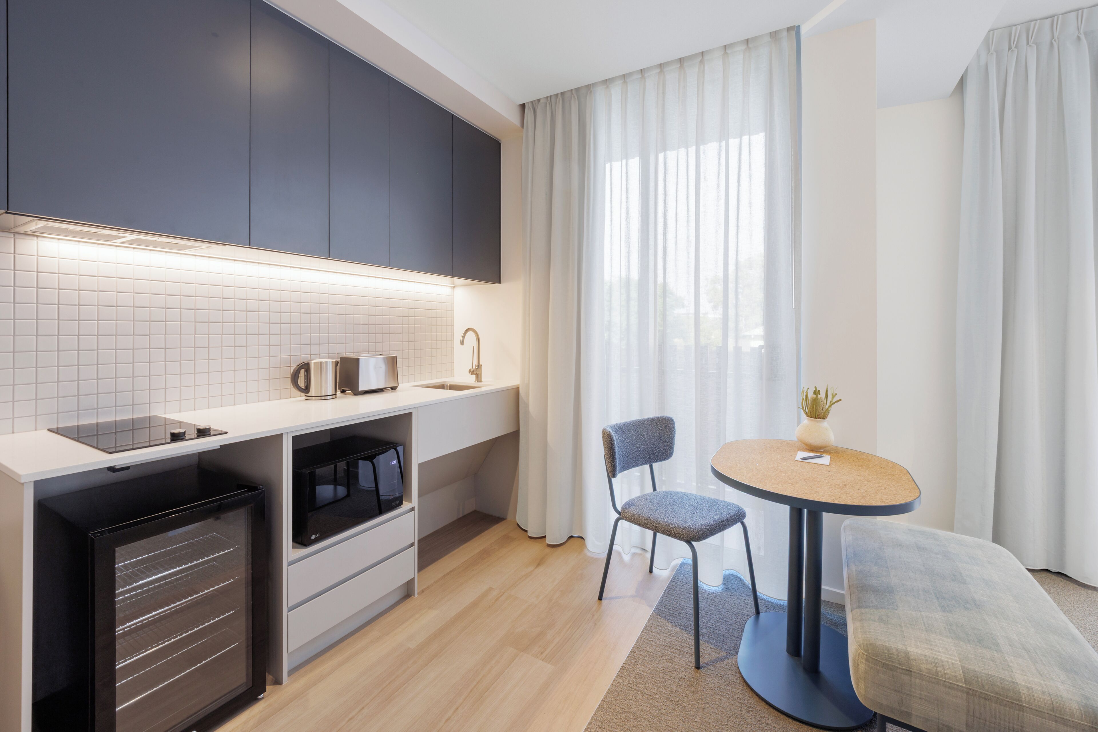 Een moderne keuken met een inbouwoven, magnetron en waterkoker, een eethoek met tafel en stoelen, en een groot raam met gordijnen.