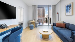 Ett modernt hotellrum med en platt-TV, en soffa, ett soffbord, ett skrivbord med en telefon och en balkong med utsikt över höga byggnader.