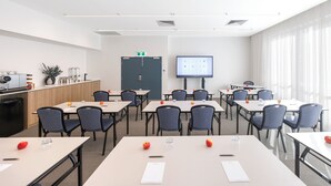 A conference room with tables, chairs, a projector screen, and a kitchenette area. - Quest Southport (Southport)