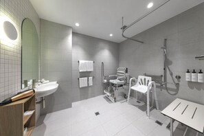A modern bathroom with a sink, mirror, shower, and a wheelchair-accessible chair. - Quest Southport (Southport)