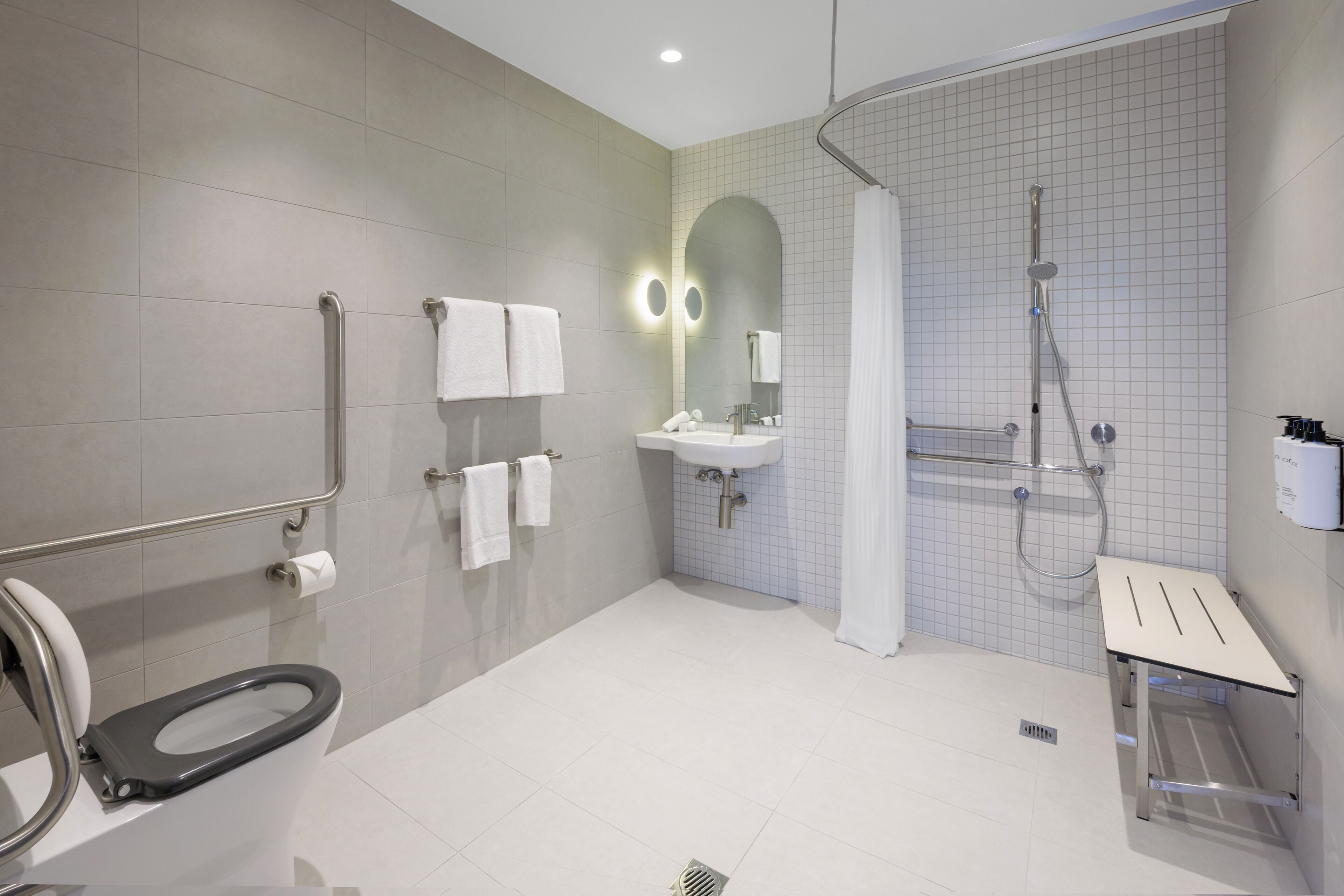 A modern bathroom with a toilet, grab bars, a sink, a shower, and a mirror.