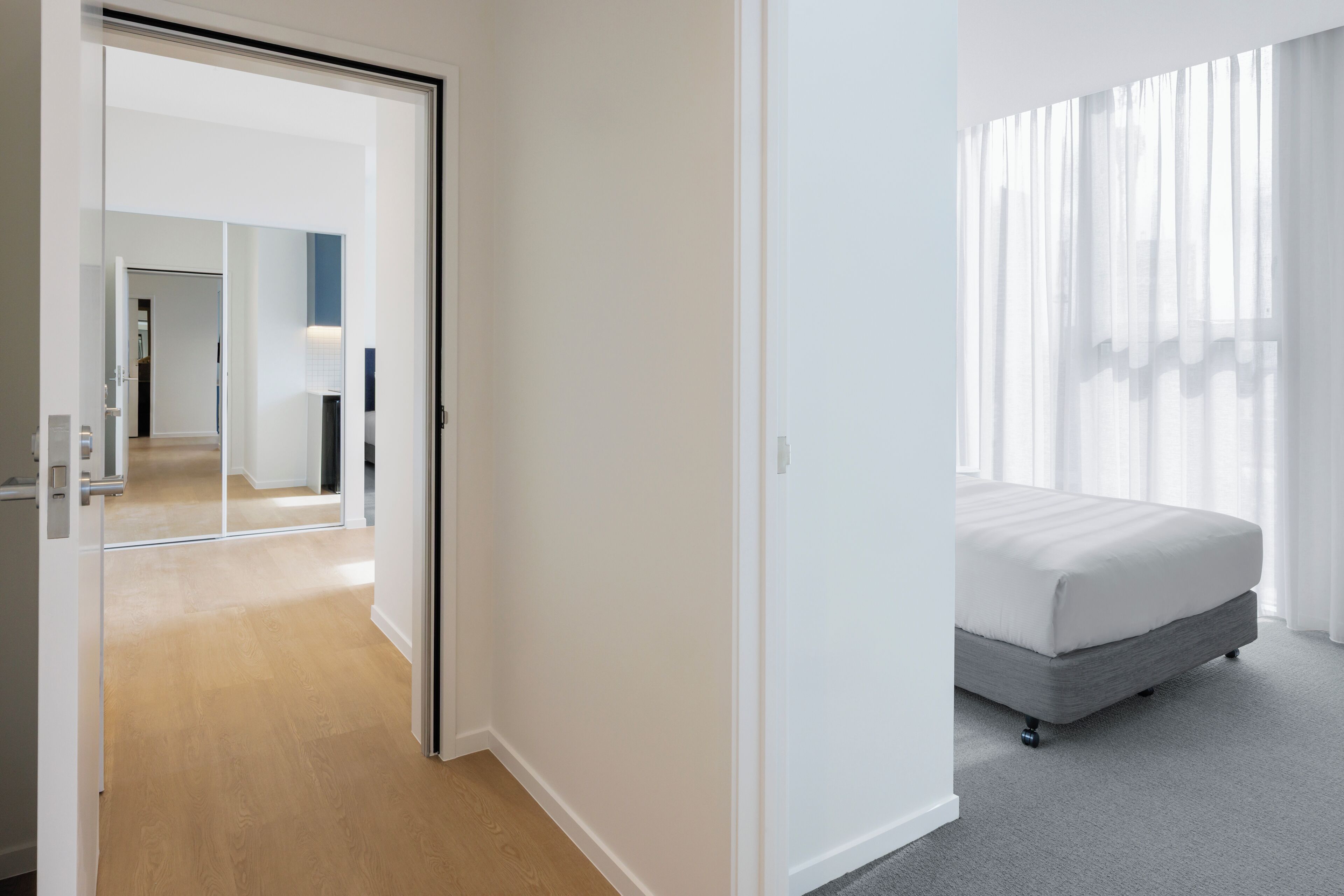 A modern hotel room with a bed, a walk-in closet, and a view of the city through sheer curtains.