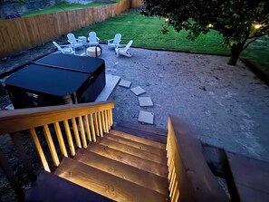 A wooden staircase leading to a backyard with a grill, white chairs, and a tree.