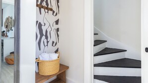 Una escalera con escalones negros y paredes blancas, un banco de madera con una cesta, y una pared con un diseño abstracto en blanco y negro.