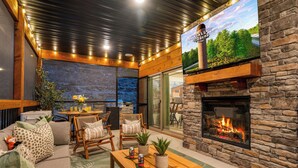 A cozy outdoor seating area with a stone fireplace, comfortable seating, and a large TV screen displaying a scenic view. - Poolside+Firepit+Hot Tub+Slides+Hidden Arcade (Hollister)