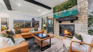 A modern living room with a stone fireplace, a large flat-screen TV, and a view of a mountain landscape. - Poolside+Firepit+Hot Tub+Slides+Hidden Arcade (Hollister)