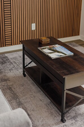 A dark wooden table with a magazine and a woven basket on top, positioned in a room with a grey carpet and wooden panel wall. - Modern 1BR w/ Massive Deck & City Views (Nashville)